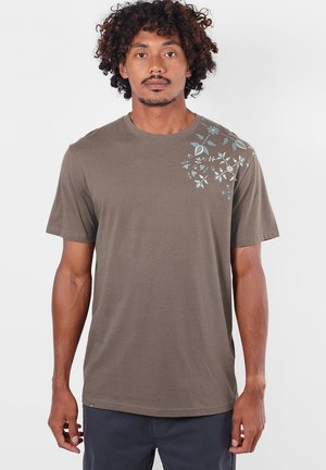 Homme aux cheveux bouclés portant un t-shirt marron à manches courtes avec des imprimés floraux bleu clair sur l'épaule gauche, debout devant un fond uni.