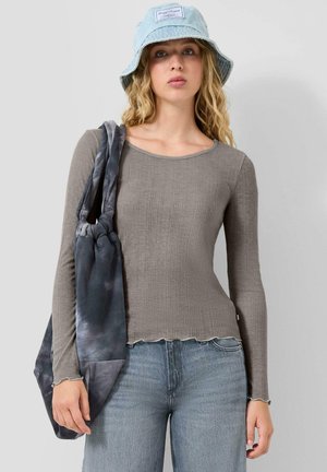 Junge Frau mit welligem Haar, die einen hellblauen Bucket-Hut, ein graues, geripptes Langarmshirt, blaue Jeans trägt und eine graue Batik-Tragetasche hält.