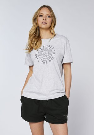 Young woman with blonde hair wearing a gray graphic t-shirt and black shorts standing with hands in pockets against a plain background.