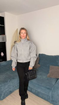 Grijze oversized sweatvest met rits en volumineuze mouwen, zwarte broek en een kleine zwarte handtas. Op de achtergrond een blauwe bank en een surfplank.