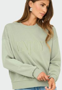 Vrouw in een lichtgroene sweatshirt met "ENJOY" geborduurd op de voorkant, die de mouwmanchet aanpast, met lang bruin haar en zilveren ringoorbellen.