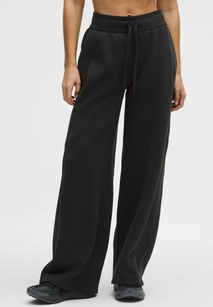 Kvinde iført sort sweatpants med højtaljet, vide ben og bindestrop i taljen samt sorte træningssko, stående mod en ensfarvet baggrund.