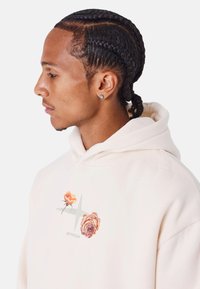 Sudadera de color marfil hecha de una tela suave, con bordados florales en naranja y rosa, con un bolsillo tipo canguro y capucha con cordón.