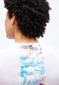 Άτομο με σγουρά μαύρα μαλλιά, φάτσα από πίσω, φοράει λευκό πουκάμισο με κάθετο πολύχρωμο αφηρημένο μοτίβο στο πάνω μέρος της πλάτης.