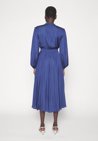 Vestido azul, de longitud hasta la rodilla, con mangas largas tipo globo, tejido texturizado, escote fruncido y una cintura ajustada. Combinado con tacones blancos.