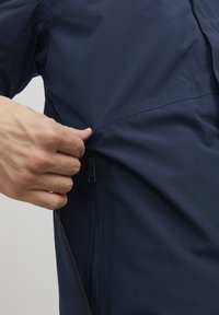 Giacca impermeabile blu navy con una texture liscia. Presenta una tasca con zip sul lato e un design elegante, che enfatizza utilità e comfort.