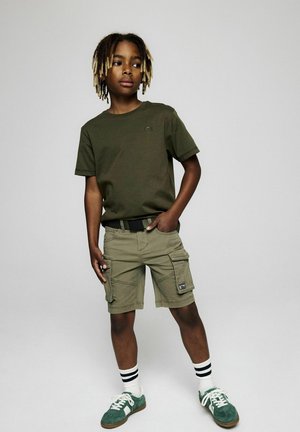 Junge mit blondspitzen Dreadlocks, der ein olivgrünes T-Shirt, Cargo-Shorts, gestreifte Socken und grüne Turnschuhe trägt und mit einer Hand in der Tasche steht.