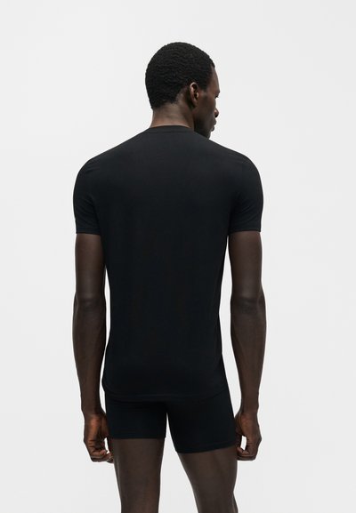 Άνδρας, ο οποίος φαίνεται από πίσω, φοράει εφαρμοστό μαύρο κοντομάνικο μπλουζάκι και μαύρο σορτς, σε απλό φωτεινό φόντο.