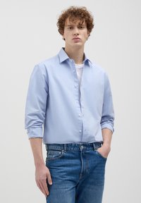 Hellblaues Hemd mit langen Ärmeln, aus Baumwollmaterial und im entspannten Schnitt. Getragen über einem weißen T-Shirt und kombiniert mit Jeans.