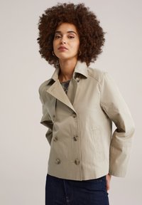 Lichtbeige, dubbelreëls jasje met een ingesneden kraag, grote knopen en schuine zijzakken. De stof heeft een gladde, licht getextureerde afwerking.