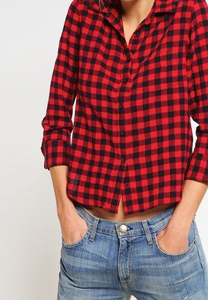 Personne portant une chemise à carreaux rouge et noir à manches longues avec poignets retroussés et un jean bleu, les mains dans les poches avant.