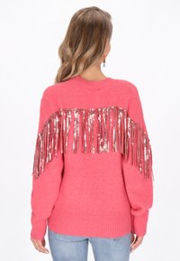 Różowy sweter z dzianiny z frędzlami i metalicznymi akcentami z tyłu. Rękawy i dół mają ściągacze, co zapewnia kontrast tekstury.