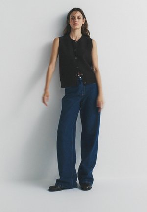 Femeie stând sprijinită de un perete alb, purtând un vestă neagră fără mâneci, cu nasturi, blugi largi de culoare albastru închis și pantofi negri.