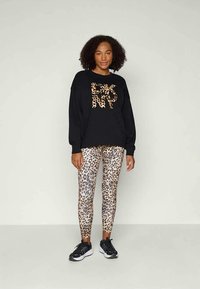 Sweatshirt preto com letras em estampado de leopardo, combinado com leggings justas também em estampado de leopardo. Usado com ténis de desporto pretos. Fundo simples.