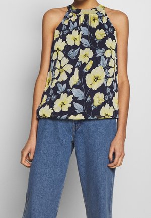 Hihaton laivastonsininen halter-toppi, jossa on suuri keltainen ja harmaa kukkakuosi, yhdistettynä sinisiin farkkuihin seisovan henkilön päällä.