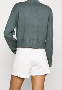 Zielony sweter dzianinowy z ribbowanym wykończeniem, połączony z białymi szortami dżinsowymi z postrzępionymi brzegami i dwoma tylnymi kieszeniami.