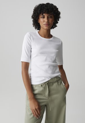 Donna con capelli ricci che indossa una maglietta bianca a maniche corte e pantaloni verde chiaro con coulisse, in piedi con una mano in tasca contro uno sfondo grigio.