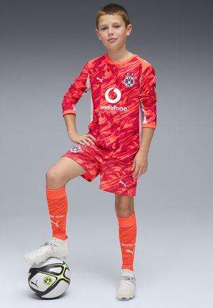 Jeugdvoetbaluniform in opvallend rood met abstract patroon, voorzien van lange mouwen, bijpassende short, oranje sokken en een wit logo.