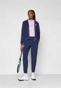 Marineblå atletisk jakke med mesh-detaljer, parret med en lys pink top og matchende marineblå joggers. Hvide sneakers med blåt accent og en tennisracket.