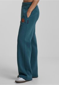 Pantalones de chándal teal con cintura elástica, bolsillos laterales y un logo bordado en naranja. La tela parece suave con una textura lisa.