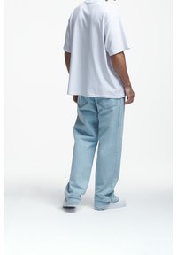 Hombre con pantalones vaqueros claros y holgados, camiseta blanca oversized y zapatillas blancas, de pie contra un fondo blanco.