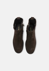 Botas de tobillo de ante marrón con una textura suave, que presentan una cremallera lateral y un diseño de punta baja y redonda. Las suelas de goma negra ofrecen agarre.