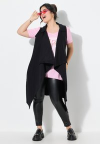 Schwarze ärmellose Weste über einem rosa Grafik-T-Shirt, kombiniert mit glänzenden schwarzen Leggings und klobigen schwarzen Schuhen. Accessoires sind pinkfarbene Sonnenbrillen.