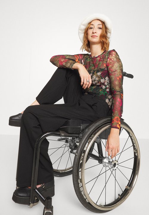 Przezroczysta bluzka z długim rękawem w kwiatowe wzory w kolorach czerwonym, zielonym i żółtym, zestawiona z luźnymi czarnymi spodniami i masywnymi czarnymi butami na platformie.