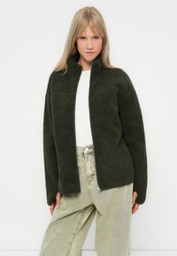 Giacca in pile verde scuro con colletto alto, taschino orizzontale sul petto e chiusura con zip. Abbinata a pantaloni larghi verde chiaro.