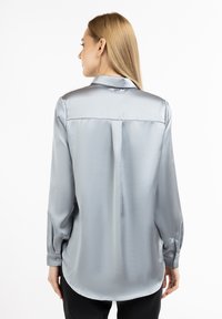 Blusa lunga grigia chiara in morbido tessuto setoso, con colletto, piega sul retro e orlo arrotondato. Polsini abbottonati e texture liscia visibile.