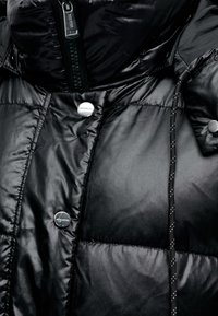 Gros plan d'une veste matelassée noire avec des boutons-pression argentés brillants, un curseur de fermeture éclair et un cordon noir avec des taches blanches.