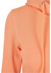 Gros plan d'un sweat à capuche de couleur pêche montrant la couture d'épaule, une partie de la manche et un cordon avec œillet métallique sur la capuche.