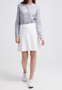 Weißer, ausgeflogener Rock kombiniert mit einer langärmeligen, blau gemusterten Bluse. Die Schuhe sind weiße schnürbare Schuhe. Glatte Texturen sind sichtbar.