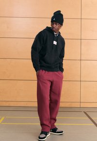 Svart hettegenser med fremre lomme, røde joggebukser og svart lue med logo. Skoene er svarte med grå detaljer og hvite såler.