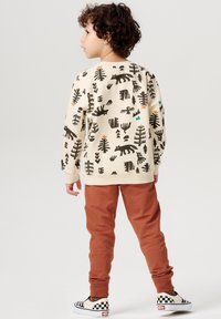 Crème sweatshirt met zwarte dieren- en bomenmotieven, gecombineerd met roestkleurige broek en geruite instappers.