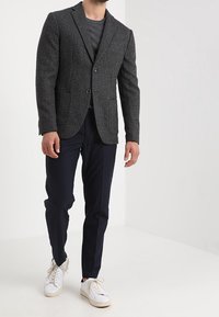 Blazer gris texturé à motif à carreaux, à deux boutons, avec un pantalon bleu marine, associé à des baskets blanches. Apparence décontractée mais soignée.