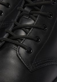 Primer plano de una bota de cuero negro con cordones redondos negros atravesados por ojales de metal y un detallado cosido.