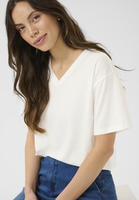 Hvid V-hals t-shirt med korte ærmer, lavet af glat stof. Modellen bærer den med blå højtaljede denim. Enkel design.