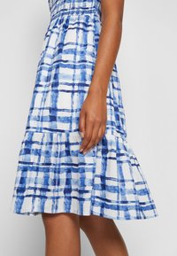 Robe à carreaux bleue et blanche longueur genou portée par une personne, montrant le milieu du corps et la main reposant à côté de l'ourlet de la robe.