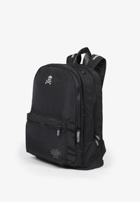 Mochila negra con un logo de calavera y tibias cruzadas, múltiples compartimentos con cremalleras, correas de hombro acolchadas y un pequeño bolsillo frontal con texto de la marca.