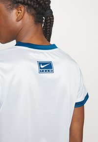 Person med flätat hår som bär en vit Nike Team sporttröja med blå detaljer, visad från ryggen och axelområdet.