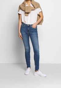 Dunne blauwe jeans gemaakt van denim, met een hoge taille en voorzakken. Gecombineerd met een wit shirt en een beige gebreide trui die over de schouders is gedrapeerd.