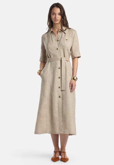 Femeie purtând o rochie cămașă bej cu nasturi, curea în talie, mâneci trei sferturi, buzunare, brățări aurii și pantofi maro cu vârf ascuțit, stând pe fundal alb.