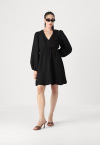 Melnas kleitas ar V veida kakla izgriezumu, savilktu jostasvietu un pūkainām piedurknēm, papildinātas ar brūniem, augstiem zābakiem ar dekoratīviem akcentiem.