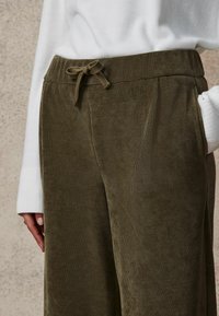 Zielone spodnie sztruksowe z elastycznym pasem i sznurkiem, z pionowym prążkowanym wzorem oraz bocznymi kieszeniami. Pas jest miękki i elastyczny.