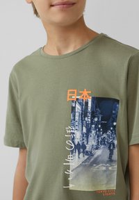 Olijfgroen katoenen t-shirt met een fotografische print van de lichten van Tokio, voorzien van oranje Japanse tekst en witte scriptaccenten.