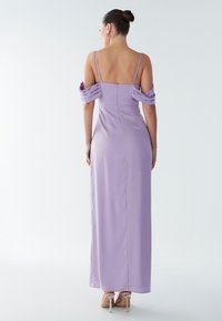 Femme portant une robe lavande longue jusqu'au sol avec des manches tombant sur les épaules et de fines bretelles, debout de dos face à la caméra.