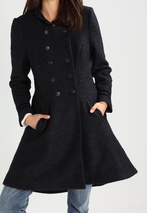 Žena v černém dvouřadém vlněném kabátu s bočními kapsami, v kombinaci se světle modrými džínami, stojící před jednobarevným pozadím.