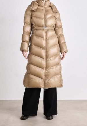 Femme portant un long manteau matelassé beige avec ceinture, debout avec un pantalon large noir et des chaussures noires sur un fond uni.
