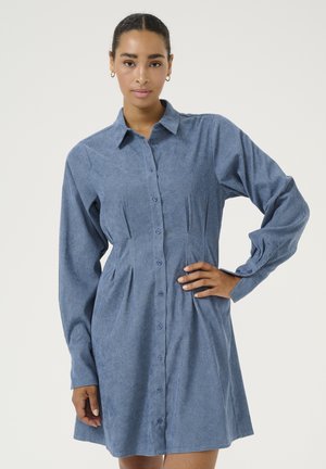 Robe chemise en tissu chambray bleu, avec des manches longues, des plis visibles et un col. Détails de boutons de couleur assortie sur toute la robe.